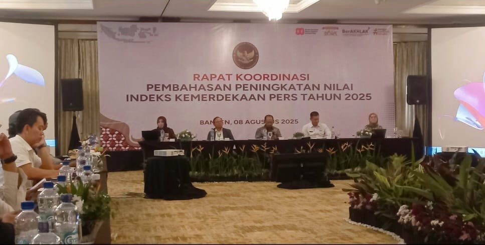 "Sebuah rapat koordinasi sedang berlangsung di ruangan konferensi dengan banner utama bertuliskan 'Rapat Koordinasi Pembahasan Peningkatan Nilai Indeks Kemerdekaan Pers Tahun 2025'. Para peserta duduk di meja panjang, siap mendengarkan presentasi atau diskusi. Di depan mereka terdapat bunga hias dan botol air minum sebagai fasilitas acara."